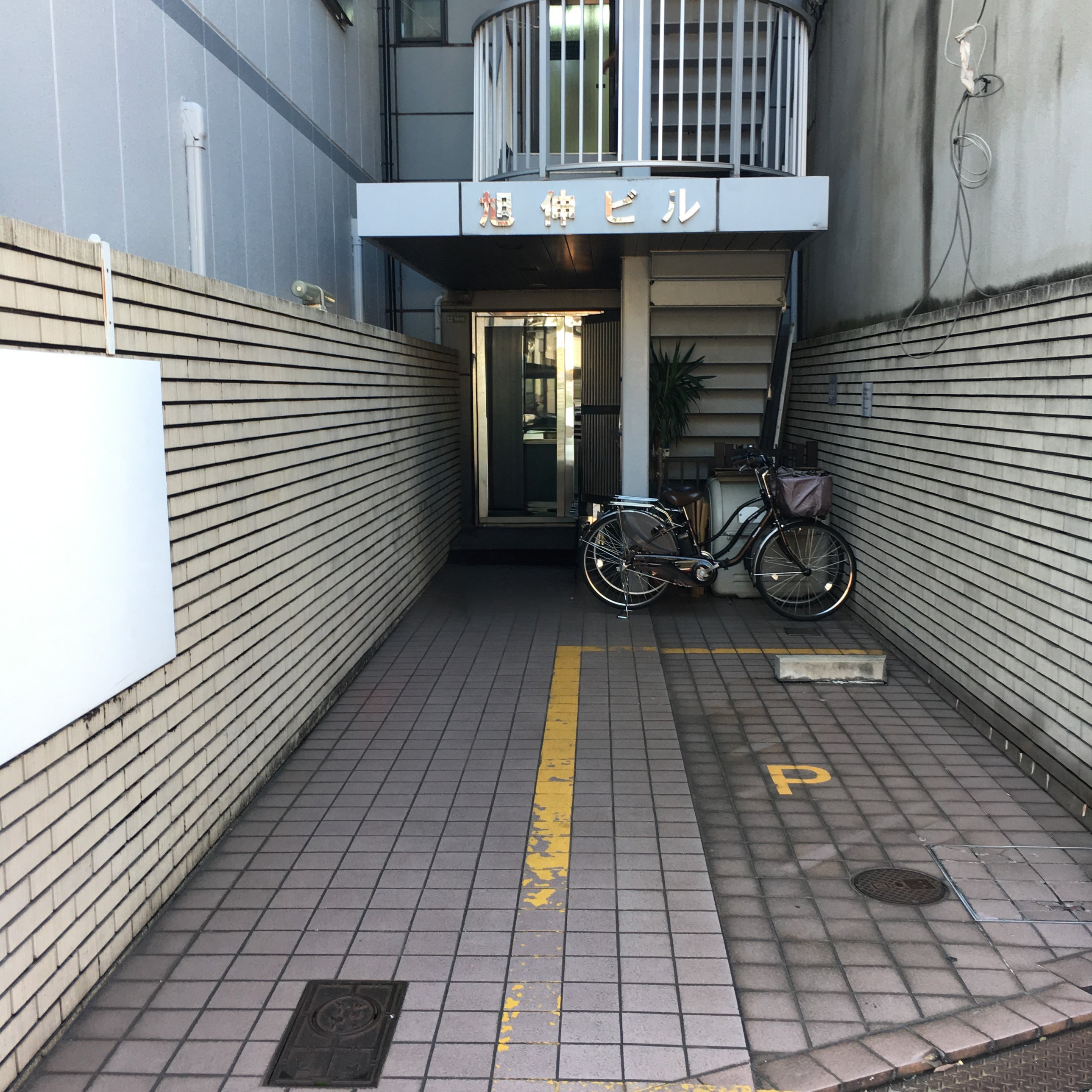 ビルの入口。左右を壁にはさまれたタイルの通路の奥に、「旭伸ビル」の看板とガラスのドアがある